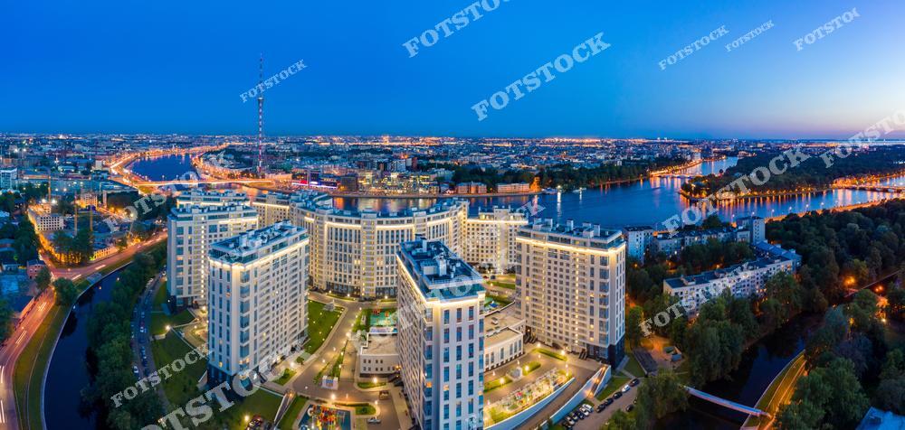 На фотографии изображён современный жилой комплекс, расположенный в центральной части Санкт-Петербурга, с панорамным видом на реку и ночные огни города. В кадре также видна Телебашня Санкт-Петербурга, являющаяся одним из заметных архитектурных элементов. Свет от зданий и улиц подчёркивает динамику и масштаб современного Петербурга. Вечерняя атмосфера передаёт гармоничное сочетание урбанистики и природы.